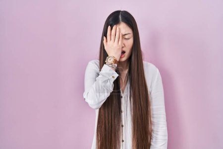 Foto de Mujer joven china de pie sobre fondo rosa bostezando cansado cubriendo media cara, ojos y boca con la mano. cara duele en el dolor. - Imagen libre de derechos