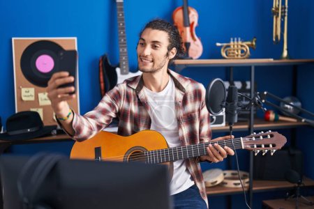 Foto de Joven músico hispano sosteniendo guitarra clásica hacer selfie por teléfono inteligente en estudio de música - Imagen libre de derechos