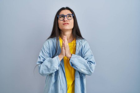 Téléchargez les photos : Young hispanic woman standing over blue background begging and praying with hands together with hope expression on face very emotional and worried. begging. - en image libre de droit