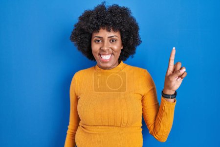 Foto de Black woman with curly hair standing over blue background showing and pointing up with finger number one while smiling confident and happy. - Imagen libre de derechos
