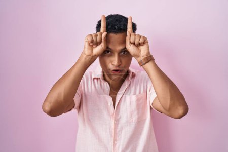 Foto de Joven hombre hispano de pie sobre fondo rosa haciendo un gesto divertido con el dedo sobre la cabeza como cuernos de toro - Imagen libre de derechos