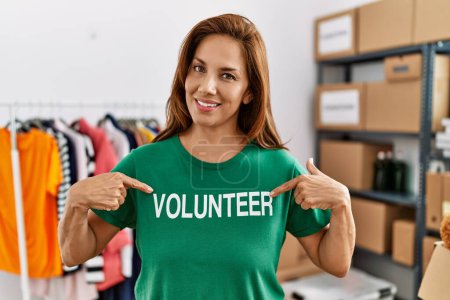 Photo for Young latin woman smiling confident pointing with fingers to volunteer uniform at charity center - Royalty Free Image