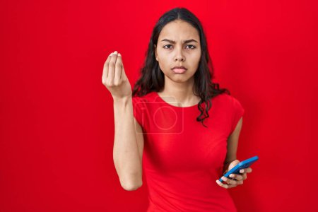 Téléchargez les photos : Jeune femme brésilienne à l'aide d'un smartphone sur fond rouge faire geste italien avec la main et les doigts expression confiante - en image libre de droit