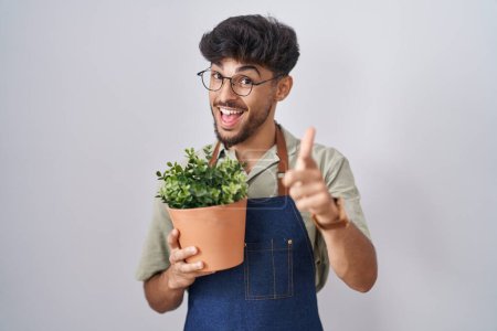 Téléchargez les photos : Arab man with beard holding green plant pot pointing fingers to camera with happy and funny face. good energy and vibes. - en image libre de droit