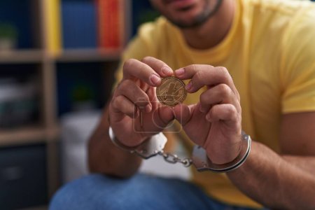 Photo for Young hispanic man criminal holding litecoin crypto currency wearing handcuffs at home - Royalty Free Image