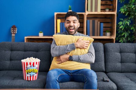 Téléchargez les photos : Homme hispanique avec barbe manger du pop-corn regarder un film à la maison souriant et riant fort à haute voix parce que drôle blague folle. - en image libre de droit