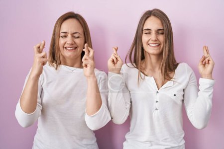 Téléchargez les photos : Middle age mother and young daughter standing over pink background gesturing finger crossed smiling with hope and eyes closed. luck and superstitious concept. - en image libre de droit