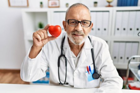 Téléchargez les photos : Homme médecin mature tenant coeur rouge à la clinique regardant positif et heureux debout et souriant avec un sourire confiant montrant les dents - en image libre de droit
