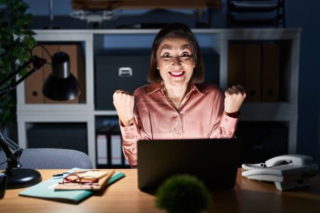 Foto de Mujer hispana de mediana edad trabajando usando computadora portátil tarde en la noche gritando orgullosa, celebrando la victoria y el éxito muy emocionada con los brazos levantados - Imagen libre de derechos