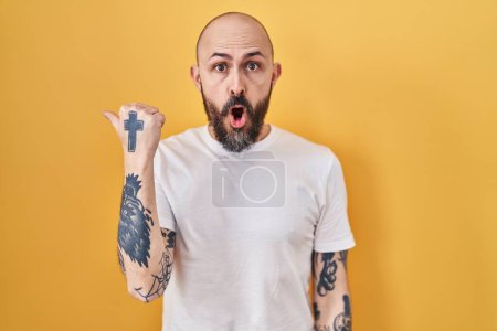 Photo for Young hispanic man with tattoos standing over yellow background surprised pointing with hand finger to the side, open mouth amazed expression. - Royalty Free Image