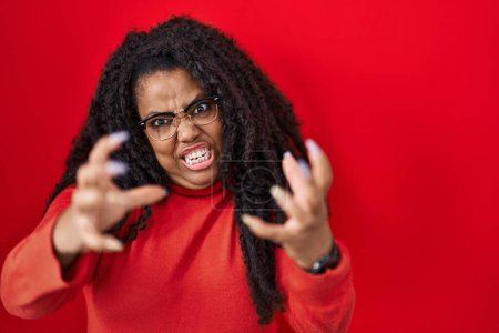 Téléchargez les photos : Plus taille hispanique femme debout sur fond rouge criant frustré par la rage, les mains essayant d'étrangler, hurlant fou - en image libre de droit