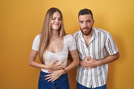 Photo for Young couple standing over yellow background with hand on stomach because indigestion, painful illness feeling unwell. ache concept. - Royalty Free Image