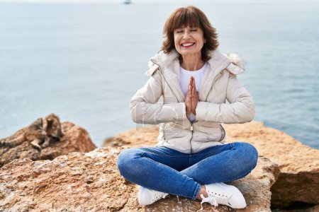 Photo for Middle age woman doing yoga exercise sitting on the rock at seaside - Royalty Free Image