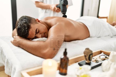 Téléchargez les photos : Jeune homme hispanique détendu ayant massage du dos à l'aide d'un pistolet à percussion au centre de beauté - en image libre de droit