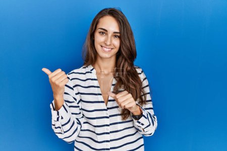 Foto de Young hispanic woman standing over blue isolated background pointing to the back behind with hand and thumbs up, smiling confident - Imagen libre de derechos