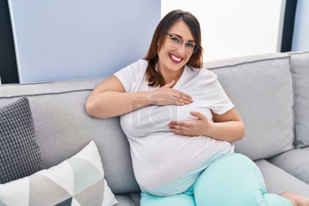 Photo for Young pregnant woman examining breast sitting on sofa at home - Royalty Free Image