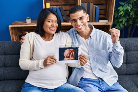 Photo for Young hispanic couple expecting a baby sitting on the sofa showing baby ultrasound screaming proud, celebrating victory and success very excited with raised arm - Royalty Free Image