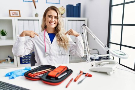 Photo for Young beautiful doctor woman with reflex hammer and medical instruments looking confident with smile on face, pointing oneself with fingers proud and happy. - Royalty Free Image