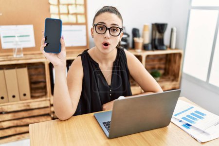 Photo for Young brunette woman holding smartphone at the office scared and amazed with open mouth for surprise, disbelief face - Royalty Free Image