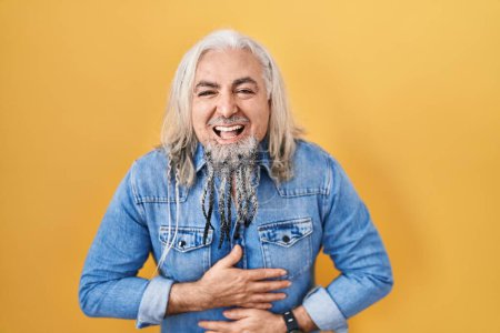 Téléchargez les photos : Homme d'âge moyen avec les cheveux gris debout sur fond jaune souriant et riant dur à haute voix parce que drôle blague folle avec les mains sur le corps. - en image libre de droit