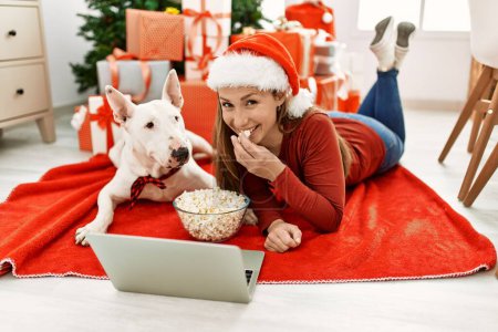 Young caucasian woman watching movie lying with dog by christmas tree at home