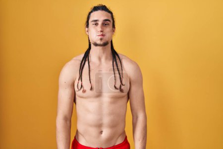 Téléchargez les photos : Hispanic man with long hair standing shirtless over yellow background puffing cheeks with funny face. mouth inflated with air, crazy expression. - en image libre de droit