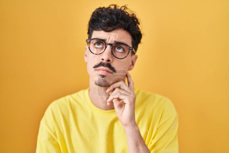 Photo for Hispanic man wearing glasses standing over yellow background with hand on chin thinking about question, pensive expression. smiling with thoughtful face. doubt concept. - Royalty Free Image