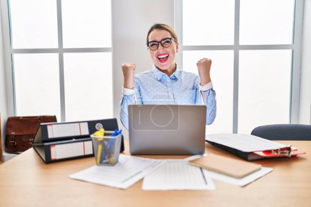 Téléchargez les photos : Young hispanic woman working at the office wearing glasses very happy and excited doing winner gesture with arms raised, smiling and screaming for success. celebration concept. - en image libre de droit
