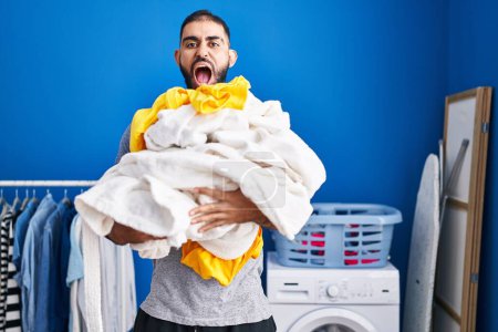 Foto de Hombre de Oriente Medio con barba sosteniendo un montón de ropa de lavandería enojado y loco gritando frustrado y furioso, gritando con ira. rabia y concepto agresivo. - Imagen libre de derechos