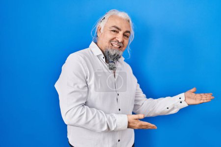Foto de Hombre de mediana edad con el pelo gris de pie sobre fondo azul invitando a entrar sonriendo natural con la mano abierta - Imagen libre de derechos