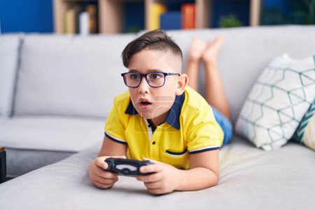 Foto de Joven niño hispano jugando videojuego sosteniendo el controlador en el sofá en la cara de choque, mirando escéptico y sarcástico, sorprendido con la boca abierta - Imagen libre de derechos