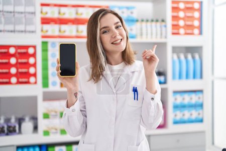 Téléchargez les photos : Blonde femme caucasienne travaillant à la pharmacie pharmacie montrant écran smartphone souriant heureux pointant avec la main et le doigt sur le côté - en image libre de droit