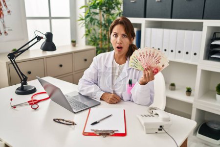 Téléchargez les photos : Hispanic doctor woman holding shekels banknotes at the clinic scared and amazed with open mouth for surprise, disbelief face - en image libre de droit