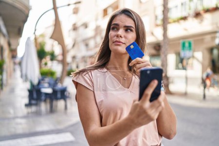 Foto de Young beautiful hispanic woman using smartphone and credit card with doubt expression at street - Imagen libre de derechos