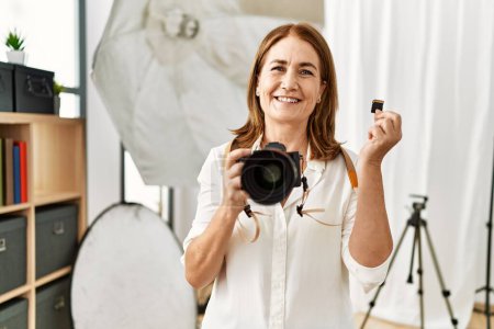 Photo for Middle age caucasian woman photographer holding professional camera and memory card at photograph studio - Royalty Free Image