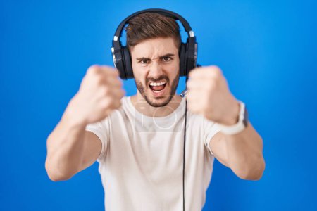 Téléchargez les photos : Hispanic man with beard listening to music wearing headphones angry and mad raising fists frustrated and furious while shouting with anger. rage and aggressive concept. - en image libre de droit
