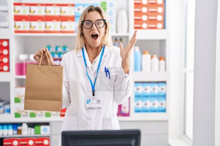 Foto de Young blonde woman working at pharmacy drugstore holding paper bag celebrating victory with happy smile and winner expression with raised hands - Imagen libre de derechos
