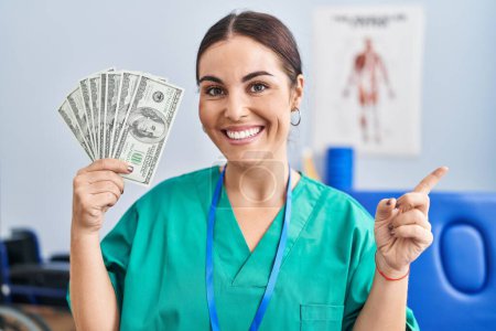 Photo for Young hispanic woman holding dollars banknotes working at pain recovery clinic smiling happy pointing with hand and finger to the side - Royalty Free Image
