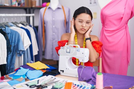 Foto de Hispanic young woman dressmaker designer using sewing machine covering ears with fingers with annoyed expression for the noise of loud music. deaf concept. - Imagen libre de derechos