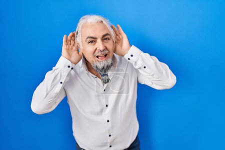 Foto de Hombre de mediana edad con el pelo gris de pie sobre el fondo azul tratando de escuchar ambas manos en el gesto de la oreja, curioso por chismes. problema auditivo, sordo - Imagen libre de derechos