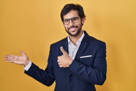Foto de Hombre latino guapo de pie sobre fondo amarillo mostrando la palma de la mano y haciendo buen gesto con los pulgares hacia arriba, sonriendo feliz y alegre - Imagen libre de derechos