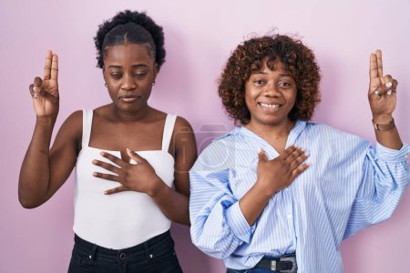 Téléchargez les photos : Deux femmes africaines debout sur fond rose souriant jurant avec la main sur la poitrine et les doigts vers le haut, faisant un serment de fidélité - en image libre de droit
