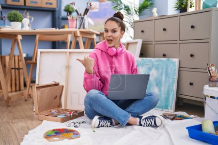 Photo for Young hispanic girl using laptop at painter studio pointing thumb up to the side smiling happy with open mouth - Royalty Free Image