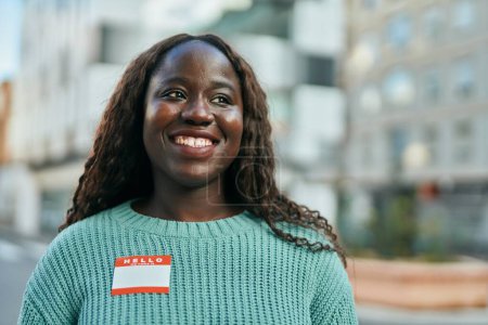 Téléchargez les photos : Jeune femme africaine souriante heureuse portant bonjour je suis autocollant à la ville - en image libre de droit