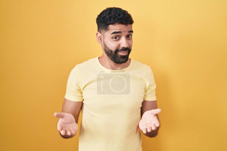 Photo for Hispanic man with beard standing over yellow background clueless and confused with open arms, no idea concept. - Royalty Free Image