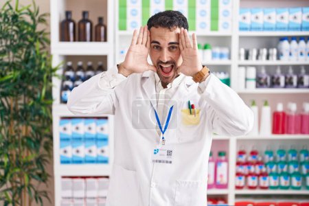 Foto de Hombre hispano guapo trabajando en farmacia sonriendo alegre jugando a echar un vistazo a un boo con las manos mostrando la cara. sorprendido y salido - Imagen libre de derechos