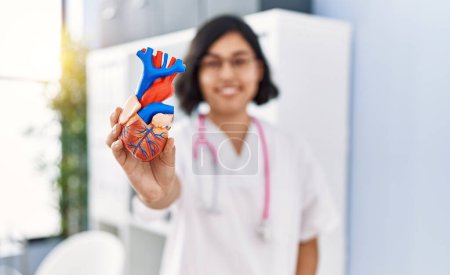 Téléchargez les photos : Jeune femme latine portant l'uniforme de médecin tenant le modèle anatomique du coeur à la clinique - en image libre de droit