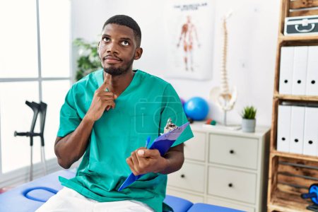 Foto de Hombre afroamericano joven que trabaja en la clínica de recuperación del dolor pensando concentrado en la duda con el dedo en la barbilla y mirando hacia arriba preguntándose - Imagen libre de derechos