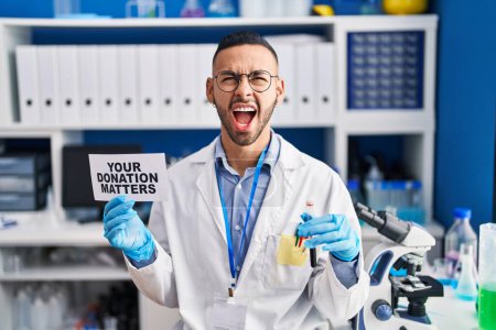 Photo for Young hispanic man working at scientist laboratory holding your donation matters holding blood sample angry and mad screaming frustrated and furious, shouting with anger looking up. - Royalty Free Image