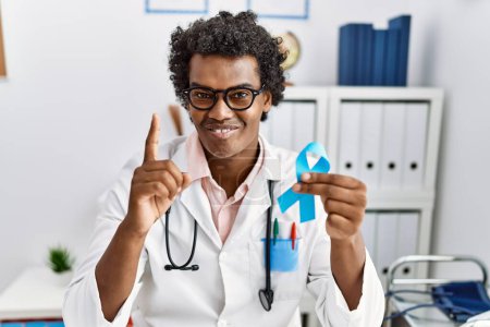 Téléchargez les photos : Docteur africain homme tenant un ruban bleu surpris par une idée ou une question pointant du doigt avec un visage heureux, numéro un - en image libre de droit